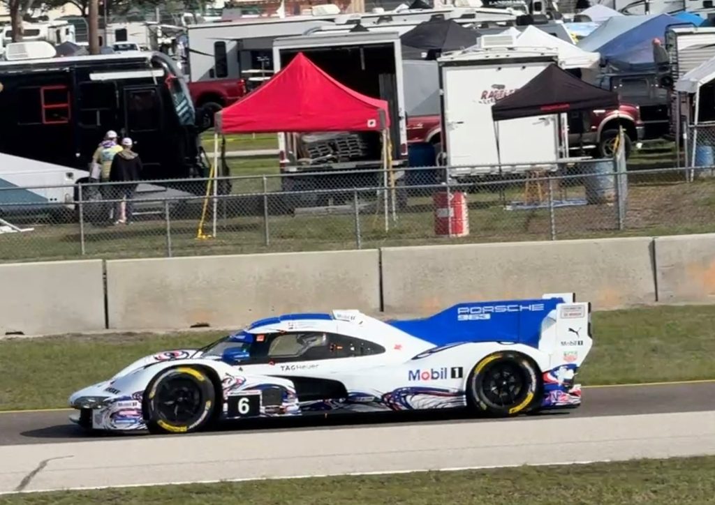 A Porsche race car numbered 6 in a blue and white design, moving on a racetrack with a grassy area and background tents and vehicles.