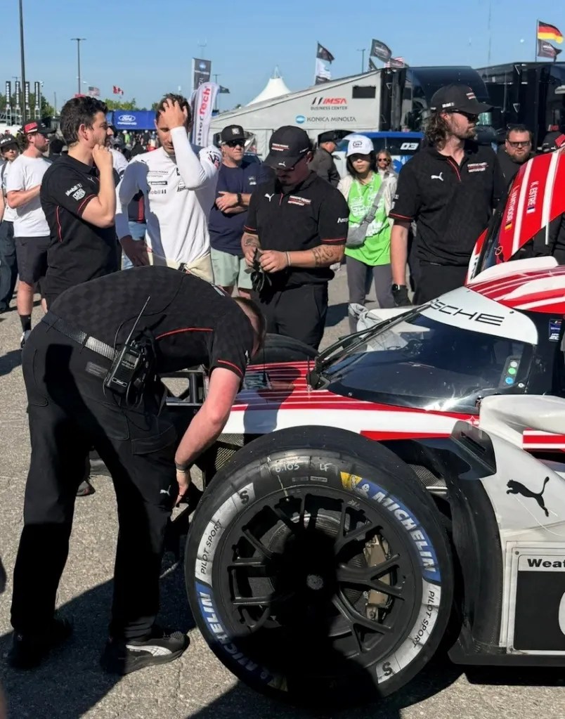 A group of people gathered around a race car, focusing on a mechanic working on the vehicle's tire. Team members in black shirts are engaged in discussions while a few onlookers observe the scene in a busy pit area.