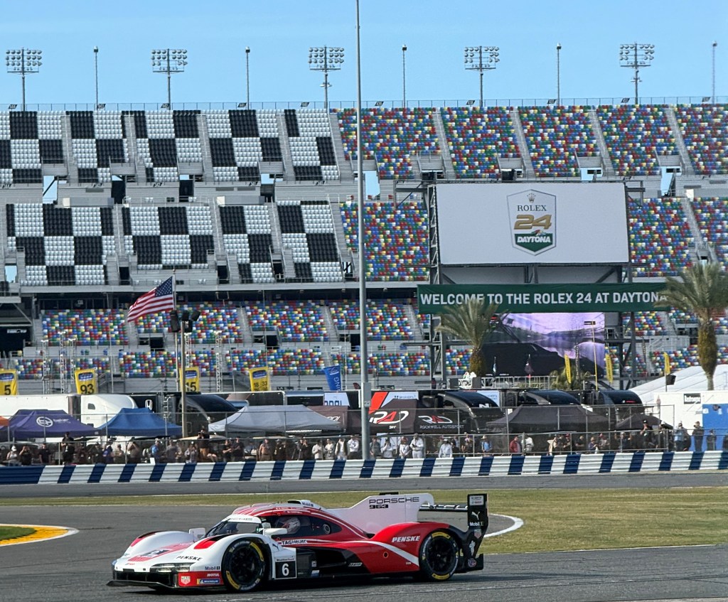 A Porsche race car undergoing a turn at the Daytona International Speedway during the Rolex 24 event, with colorful grandstands and a large electronic sign in the background.
