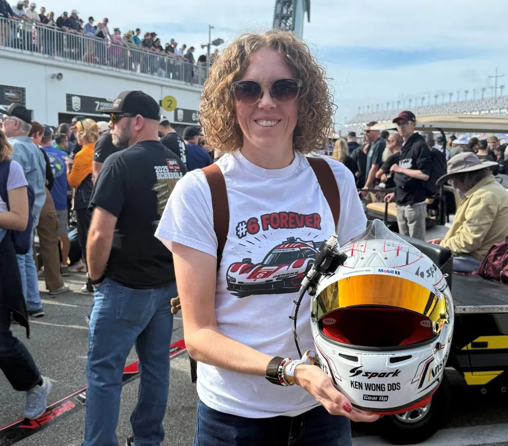 A woman with curly hair and sunglasses smiles while holding a racing helmet at a motorsport event, surrounded by a crowd.