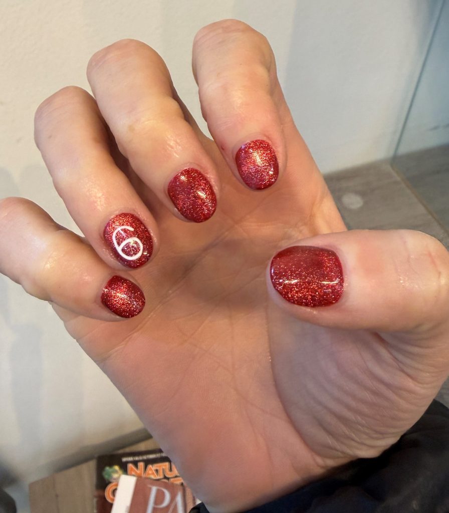 A hand displaying manicured nails painted with red glitter polish, featuring a small white design on one nail.