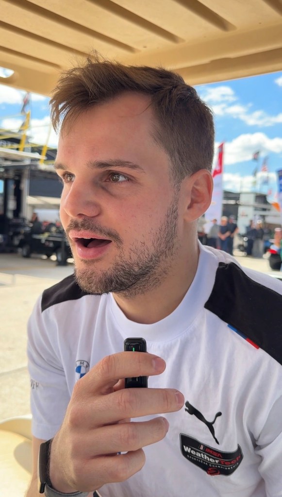 A young man with short hair and a slight beard, wearing a white shirt with a logo, appears surprised while speaking into a microphone. The background includes a clear blue sky and blurred figures, suggesting an outdoor event.