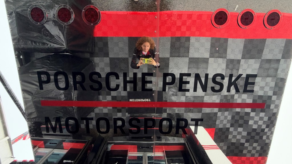 Reflection of a person holding a phone in front of a Porsche Penske Motorsport sign with a checkered background.