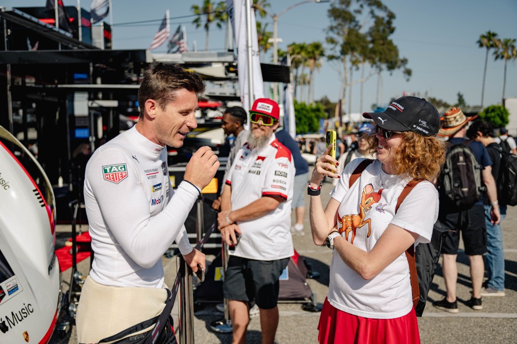 A man in a racing suit speaking to a woman holding a phone; the woman is wearing a t-shirt featuring a deer and a red skirt, while another man with a white beard in a racing shirt stands nearby.