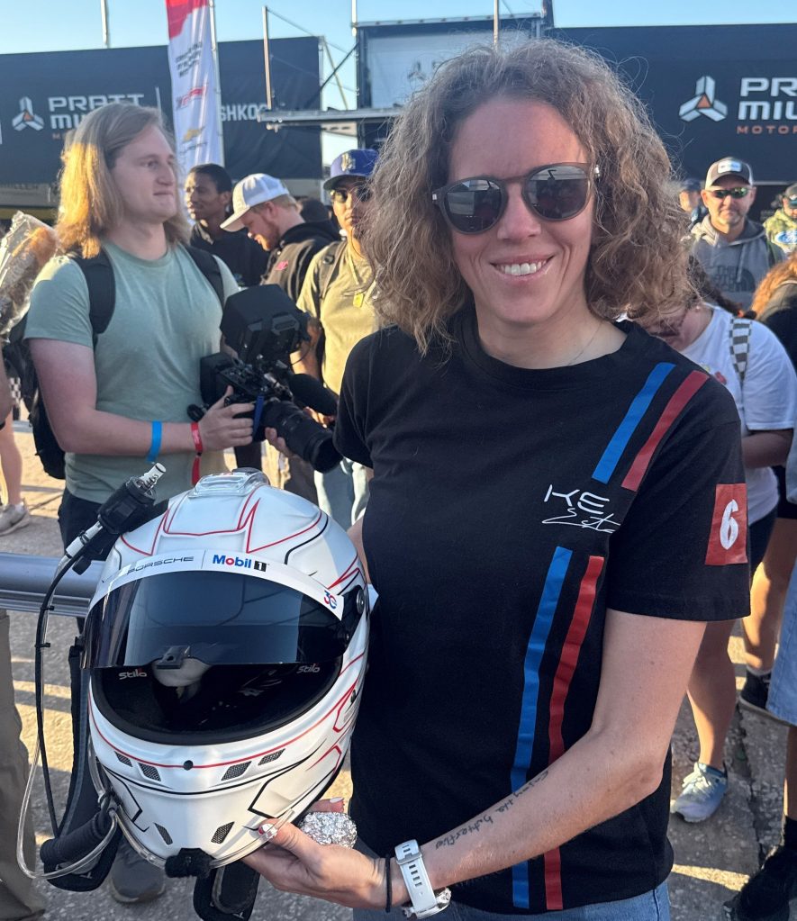 A woman with curly hair wearing sunglasses holds a racing helmet with a white base and red accents, standing in a crowd at an event.