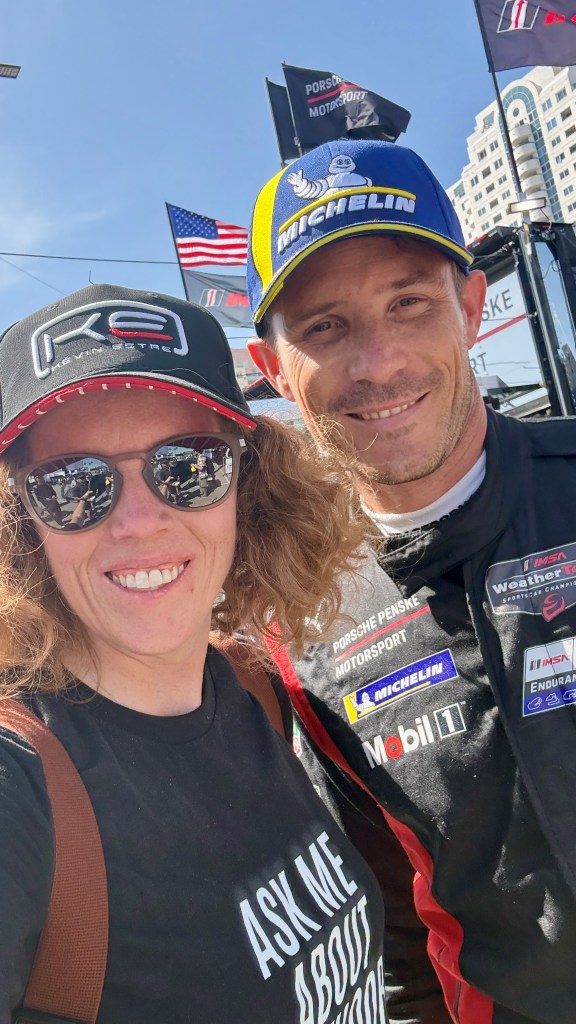 A woman and a race car driver pose for a selfie, both smiling. The woman wears a black shirt with the text 'ASK ME ABOUT KARTING' and a cap, while the driver sports a blue and yellow branded cap and a racing suit. In the background, there are flags and an urban setting.