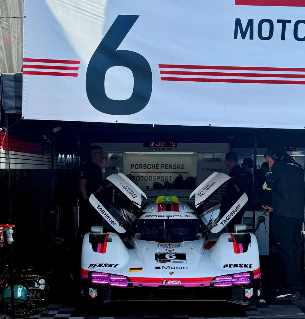 A Porsche race car with the number 6 displayed, positioned in a pit area with crew members working around it. The car features a sleek design with distinct branding and raised rear wing.