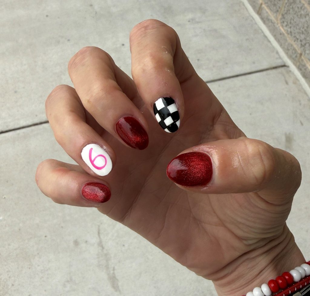 Close-up of a hand showing manicured nails, featuring red glitter polish on most nails, a white nail with a pink number '6', and one black and white checkered nail.