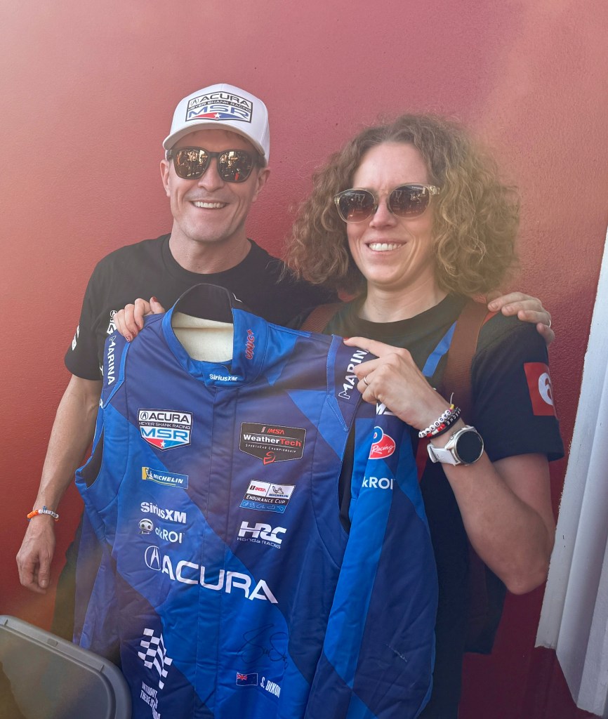 Two people smiling while holding a blue racing suit with various sponsor logos, standing against a red background.