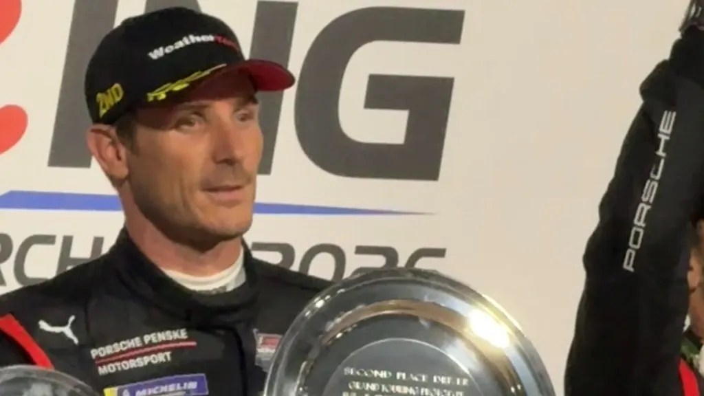 Racing driver celebrating with a trophy, wearing a black racing suit and cap with the number '2nd' visible.